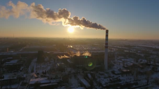 Aerial shot of a towering hot gas chimney with white smoke at sunset in winter A bird`s eye view of a sky- high chimney with a dark smoke stream at a splendid sunset in winter. The cityscape is industrial and snowy — Stock Footage Aerial Shot Towering Hot Gas Chimney White Smoke Sunset Winter Stock Video