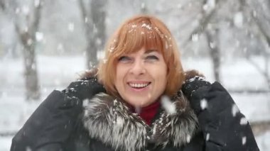 Yağan kar ve onun hood slo-mo bir parkta ağır kar taneleri altında kışın gülümseyen sarışın bir kadın harika bir manzarasını kalkış altında bir neşeli kadın gülümsüyor. O onun kürk dururlar ve elleri slo-mo yükseltir