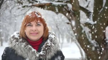 Orta yaşlı sarışın bir kadın duruyor ve gülümsüyor kış bir parkta kar taneleri altında bir kürk mutlu sarışın kadın iyimser bir bakış ceket. O neşeyle büyük bir ağaç ve harika bir parkta ağır kar taneleri altında kışın gülümsüyor. 