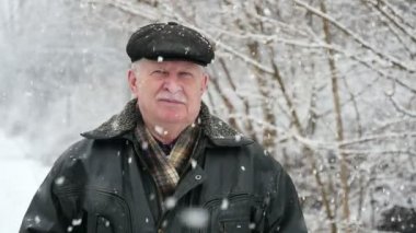 Yaşlı, bilge bir adam görkemli bir parkta beyaz kar taneleri altında bir üst düzey gri saçlı adam bir ağır ceket ve düz bir kapak sanat konusunda hava atan bir görünümünü kışın duruyor. Kışın karlı bir parkta duruyor. Kar taneleri üstünden Jakuzziler.