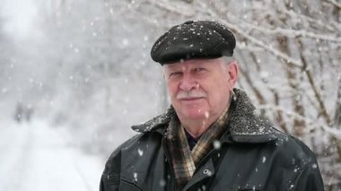 Mutlu bir yaşlı adam görkemli bir parkta ağır kar taneleri altında beyaz mustashe düz bir kapak ile üst düzey bir adam inanılmaz bir görünümünü kışın duruyor. O gülümser ve kışın karlı bir parkta duruyor. Kar taneleri üstünden Jakuzziler.