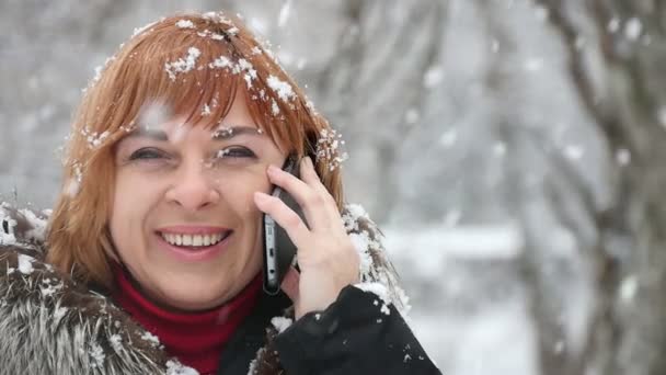 一个快活的女人在一个手机上说话 在公园 Falliing 的雪花下微笑 看到一个穿着毛皮大衣的金发女郎的笑脸 她在冬天的一个公园里 在空中飞舞的雪花下谈论移动 她感到高兴 — 图库视频影像