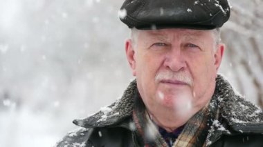 Yaşlı, bilge bir adam güzel bir parkta ağır kar taneleri altında kışın cesur kıdemli biri beyaz mustashe düz bir kapak ile etkileyici bir manzara anlamına gelir. Kışın karlı bir parkta duruyor. Kar taneleri üstünden spin.