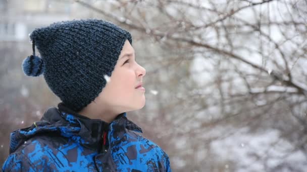一个快乐的男孩在小巷里看着 飘落的雪花在冬天是美妙的 一个九岁男孩戴着黑色针织帽子的令人鼓舞的特写镜头 他在冬天的小巷里看着魔术和奇妙的雪花 — 图库视频影像