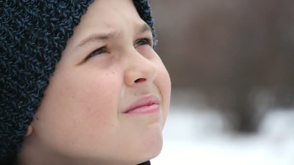 一个小男孩在小巷里看着 飘落的雪花在冬天是美丽的 一个九岁男孩戴着黑色针织帽子的特写镜头 他在冬天的一个艺术小巷里看着大而美丽的雪花 — 图库视频影像