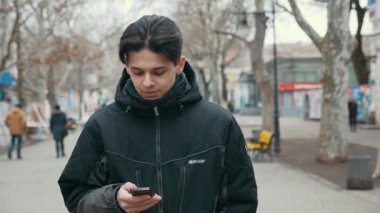 Stylisn genç bir adam bir sokak yürür ve cep telefonunu çekimde yavaş kışın park sokak strolling siyah bir parka bir genç adamın heyecan verici bir görünüm. O derinden onun düşüncelerinde gömülür.