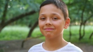 Küçük bir çocuk gülümsüyor ve yeşil bir parkta bir yol üzerinde slo-mo gün batımında yavaş yaz aylarında muhteşem bir parkta duran bir düşünceli dokuz yıl çocuk bir closeup duruyor. O düşünceli ve iyimser görünüyor
