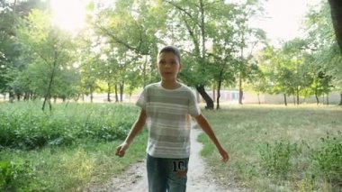 Küçük bir çocuk boyunca yeşil bir park lane günbatımı bir sportif dokuz yıl çocuğun ileride park yaz yavaş seyir bir sihirli yürüyüş yavaş hareket A muhteşem görünümünde dolaşır. O çizgili bir tshirt ve kot pantolon.