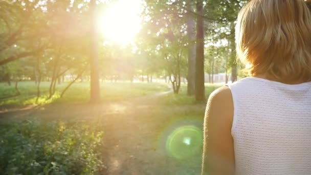 一个苗条的金发女郎沿着一条小路朝着灿烂的日出在公园里一个美丽的女人在一条无袖的白色连衣裙上 在夏天在斯洛伐克的公园里 在一幅风景如画的日出中动人的阳光 — 图库视频影像
