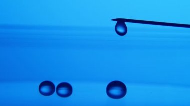 Parlayan su damlaları metalik bir iğneden bir laboratuvar mavi yüzey üzerinde büyük bir parlayan damla ve küçük BLOB'lar üzerinde bir laboratuvar mavi arka plan metalik bir iğne üzerinden düşen bir sanatsal makro çekim aşağı gel