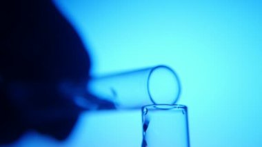 Karanlık el tüp bebek tutar ve etkileyici bir manzara mavi arka planda başka bir içine bir tüp akan bir şeffaf sıvı tıbbi bir laboratuvarda bir tane içine sıvı dökülen. Bazı karanlık ellerini tutar bir tüp.