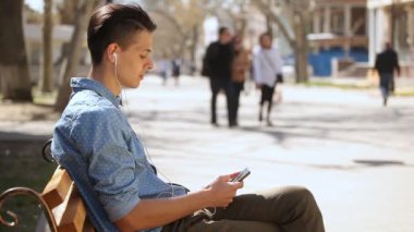 Genç adam bahar profil sportif genç bir adam onun smartphone kullanarak ve bahar güneşli bir günde harika bir arka sokakta bir bankta oturmuş bir kolsuz tişörtlü bir sokak bankta oturan onun smartphone ekran dokunur