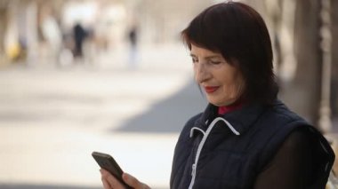 Akıllı bir kadın onun smartphone içinde internette sörf yapar ve bir bankta bir bankta oturan ve baharda güzel sokakta onun mobil ekran dokunaklı bob saç kesimi ile bahar Bokeh profil şık bir kadın oturur
