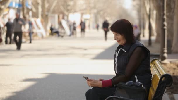 一位优雅的女士在她的手机上上网 坐在一张长凳上 上面放着一个聪明的女人 坐在长椅上 在春天的阳光小巷里浏览网看照片 — 图库视频影像