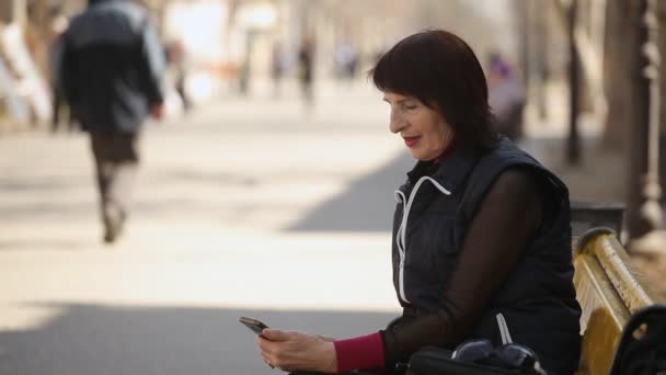 中年の女性彼女の携帯電話でネットにログインし ベンチに座っていると 春に見事な路地でいくつかの情報を得るためにネットをサーフィンにボブのヘアカットと春流行の女性のプロファイルのベンチに座っています — ストック動画