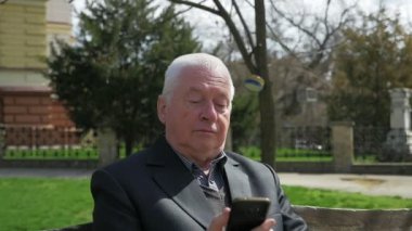 Komuta sizde oturur ve onun Smartphone cep telefonunu internette gezinme, onun hayattan zevk ve baharda bir arka sokakta bir bankta oturmuş ak başlı bir adamın bahar A 180 derece çekimde bir arka sokakta net sörf