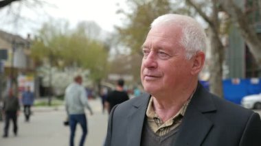 Deneyimli adam bahar yavaş kendinden emin bir ak başlı adam gülümseyen ve ağaçlar ve modern bir sokakta bir bankta oturmuş bir profil slo-mo bahar evleri sokak şehirde bir bankta oturur 