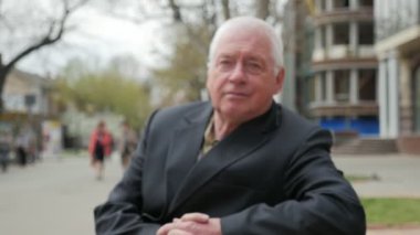 Bilge adam oturur bir şehirde sokak bahar bahar slo-mo çıplak ağaçlar ile sokak ve modern bir şehirde bir bankta oturan emin ak başlı adam görüntüsünü yakınlaştırmak evler yavaş çekimde bir bankta yaşlı 