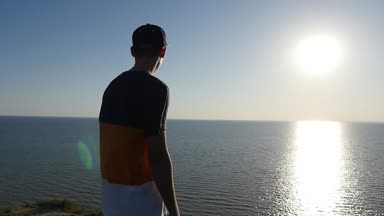 Zappy genç adam slo-mo arka görünümünde çizgili tişört, kap ve Karadeniz ayakta ve slo-mo gün batımında sihirli güneş yol zevk pantolon iyimser bir adamın parlayan batımında Karadeniz kıyısında duruyor