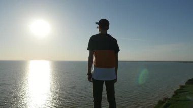 Cesur genç adam neşeli bir adamın çizgili tişört, şapka ve Karadeniz ayakta ve slo-mo gün batımında muhteşem güneş yol zevk pantolon slo-mo arka görünümünde parlayan batımında Karadeniz kıyısında duruyor