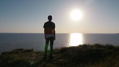 Rüya genç adam slo-mo arka görünümünde çizgili tişört ve Karadeniz kıyısında ayakta ve slo-mo gün batımında parıldayan güneş yol zevk pantolon içinde mutlu bir adamın köpüklü batımında Karadeniz kıyısında duruyor