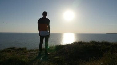 Karadeniz sahilinde slo-mo arka görünümünde bir beyzbol şapkası ve Karadeniz kıyısında ayakta ve slo-mo gün batımında altın güneş yol zevk bir T-shirt sportif bir adamın parlayan batımında romantik genç adam standları