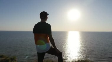 Çabuk genç adam bir mürettebat saç kesimi kap ve Karadeniz kıyısında ayakta ve tek bacak slo-mo iyi batımında bükük tutmak bir T-shirt ile sportif bir adamın slo-mo profilde altın batımında Karadeniz kıyısında duruyor