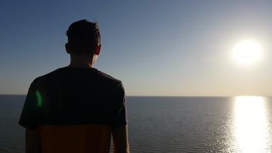Sportif genç adam bir rüya genç adamın slo-mo arka görünümünde dalgalar bir T-shirt Karadeniz sahilinde ayakta ve slo-mo altın güneş yol zevk içinde mürettebat saç kesimi ile bakar, Karadeniz kıyısında duruyor