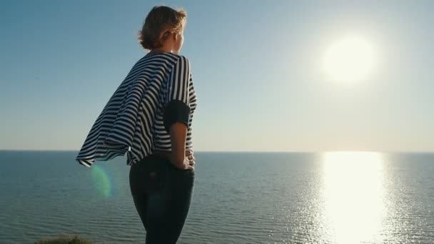 スローモーションで太陽パスと素敵な夕日でスローモーション黒海のビーチに手を振っているストライプ ブラウス立って華やかな細い女性の背面ビューでサンセットに黒海の海岸に立っている詩的なブロンド女性 — ストック動画