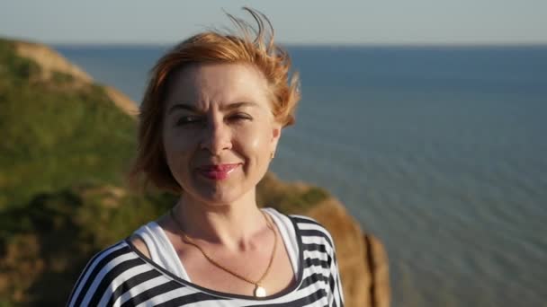 快活的妇女站立在黑海海岸和微笑在夏天在斯洛伐克特写一个滑稽的金发妇女在一条条纹的海衬衣保持手在她的腰部和微笑在黑海在夏天在日落斯洛伐克 — 图库视频影像
