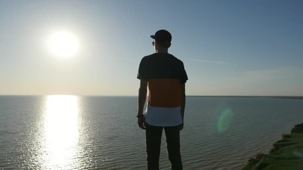 スローモーション バック ストライプの シャツ キャップ パンツ黒海に立って スローモーションで日没で立派な太陽パスを楽しんでで陽気な人間観に輝く夕焼けに黒海の海岸に立っている勇敢な若い男 — ストック動画