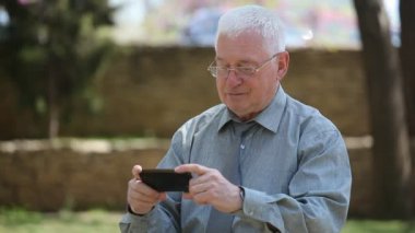 Neşeli yaşlı adam oturur ve onun smartphonescreen bir sokakta güneşli bir günde bir gri gömlek ve kravat gülümseyen ve yaz aylarında onun smartphone bir sokakta neşeli bazı video izlerken neşeli yaşlı erkeklerde etkileyici bir manzara görünüyor