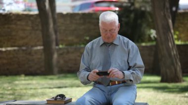 Gri saçlı adam oturur ve onun telefon ekranında bir caddede güneşli bir günde bir heyecan verici bir gri gömlek ve kravat gülümseyen ve yaz aylarında telefonunu bir sokakta neşeli bazı video izlerken iyimser bir yaşlı adamın görünüm