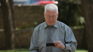 Beyaz saçlı adam duruyor ve onun smartphone ekranında bir caddede yaz aylarında bir gri gömlek ve kravat gülümseyen ve yaz aylarında onun smartphone bir sokakta bazı komik video izlerken neşeli yaşlı erkeklerde iyimser bir bakış görünüyor