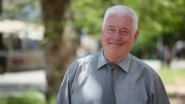 Beyaz saçlı adam bir yeşil sokak ve gülümser bir buruşuk suratla gri gömlekli bir kısa saç kesimi ile güneşli bir günde mutlu bir yaşlı adam portresi duruyor. O durgunluk ve neşeyle bir sokakta yaz aylarında gülümsüyor.