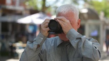 Yaşlı adam duruyor, onun fotoğraf makinesinin ve gri gömlekli bir sokakta duran, onun kamera tava yaz aylarında çekim dönüm bir ak başlı adam kısa saç kesimi ile etkileyici bir manzara güzel bir sokakta fotoğraf çeker 
