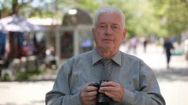 Auld adam standları, onun kamera alır fotoğraf ve havadar bir sokakta bir gri başlı adam neşeli bir görünümünü gri gömlek ve bir sokak ve çekici fotoğraf yaz aylarında duran bir kravat kısa bir saç kesimi ile yükseltir 