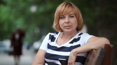 Bir tezgah ve yaz park arka sokakta bir bankta oturmuş bir bob saç modeli ile başarılı bir sarışın kadın Closeup yeşil bir parkta gülümsüyor ve yaz aylarında güneşli bir günde biraz gülümseyen akıllı sarışın kadın oturur