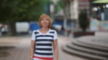 Güneşli bir günde çizgili bir bluz ve bir kameraman için yürüyüş ve mutlu bir yeşil yaz aylarında güneşli bir sokak gülümseyen kot neşeli sarışın bir kadın portresi mutlu sarışın kadın gülümser ve yeşil bir sokak gider