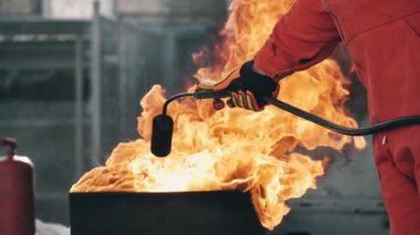 Üniformalı bir itfaiyeci, alev makinesini yanan bir küvetin üzerinde tutarak ateş püskürtüyor. Alev makineli bir itfaiyeci ve büyük bir hortum onu alev saçan bir tencerenin üzerinde tutarak sihirli bir şekilde dans ediyor.