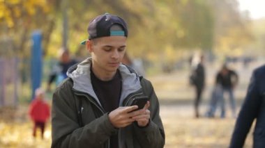 Zeki bir genç adam bir ara sokakta duruyor ve ağları tarıyor. Yavaş çekimde. Şapkalı neşeli bir genç adam ve güzel bir parkta turuncu kaplı topraklarla duruyor ve yazın telefonunda yavaş çekimde bilgi arıyor.