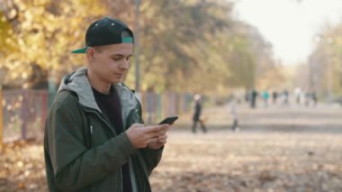 Parkta duran ve sonbaharda ağlara yavaş çekimde göz atan zeki bir genç adam. Şapkalı ve ceketli zeki bir gencin resimli bir parkta durup toprağı paslı yapraklarla kapladığı ve yazın ağır çekimde internette gezindiği heyecanlı bir profil.