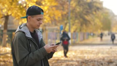 Neşeli genç adam bir ara sokakta duruyor ve sonbaharda internette yavaş çekimde sörf yapıyor. Şapkalı ve ceketli bir gencin etkileyici bir profili var. Parkta duruyor ve güneşli bir yaz gününde yavaş çekimde internette geziniyor..