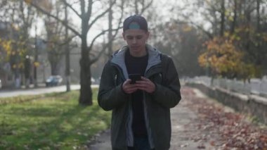 Parkta yürüyen ve sonbaharda ağda ağır çekimde sörf yapan zeki bir genç adam. Parkta taş bir korkuluk boyunca yürüyen ve güneşli bir yaz gününde ağda gezinen şapkalı ve ceketli zeki bir genç.