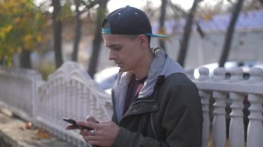 Şapkalı, kapüşonlu, taş bir korkulukta oturan ve sonbaharda yaprakları dökülen bir parkta telefonuyla internette sörf yapan zeki bir genç adamın muhteşem manzarası.