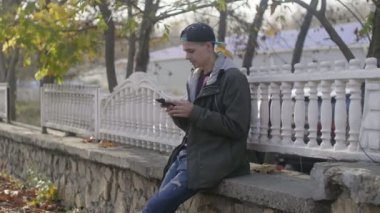 Zeki bir genç adam, sonbaharda taştan bir korkulukta oturup internette geziniyor. Şapkalı, kapüşonlu, zeki, genç bir adam, taş bir korkulukta oturuyor ve sonbaharda dökülen yapraklarla parktaki telefonunda bilgi arıyor.. 