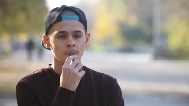 Νεαρός Άνδρας Στέκεται Και Καπνίζει Ηλεκτρονικό Τσιγάρο Ένα Γραφικό Πάρκο — Αρχείο Βίντεο