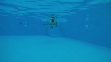 Suyun altında yüzen maskeli neşeli çocuk yavaş çekimde yüzen bir havuzda. 11 yaşında şort giymiş küçük bir esmer çocuk ve güneşli bir yaz gününde, güneşli bir havuzda yüzen maske takan bir kurbağacık..