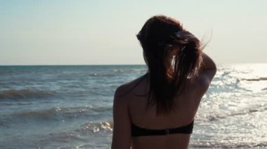 Neşeli genç bir kadın yaz aylarında uzun saçlarıyla kıyı şeridinde ağır çekimde oynar. Bikinili güzel bir esmer kadının güneşli bir yaz gününde kumsalda uzun saçlarıyla el sallaması ve oynaması inanılmaz..