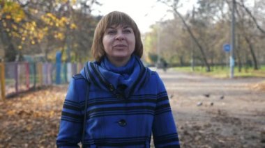 Sonbaharda güneşli bir parkta gezinen ve gülümseyen mutlu sarışın kadın. Kısa saçlı neşeli sarışın bir kadın. Şık ceketli, güneşli bir parkta ağır çekimde geziniyor ve gülümsüyor.